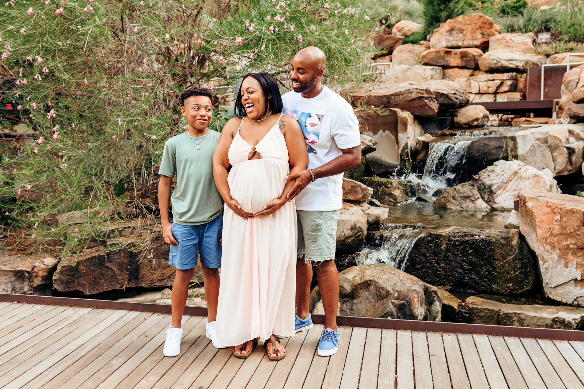 Family of 3 laugh in front of waterfall at Myriad Gardens during maternity photoshoot.